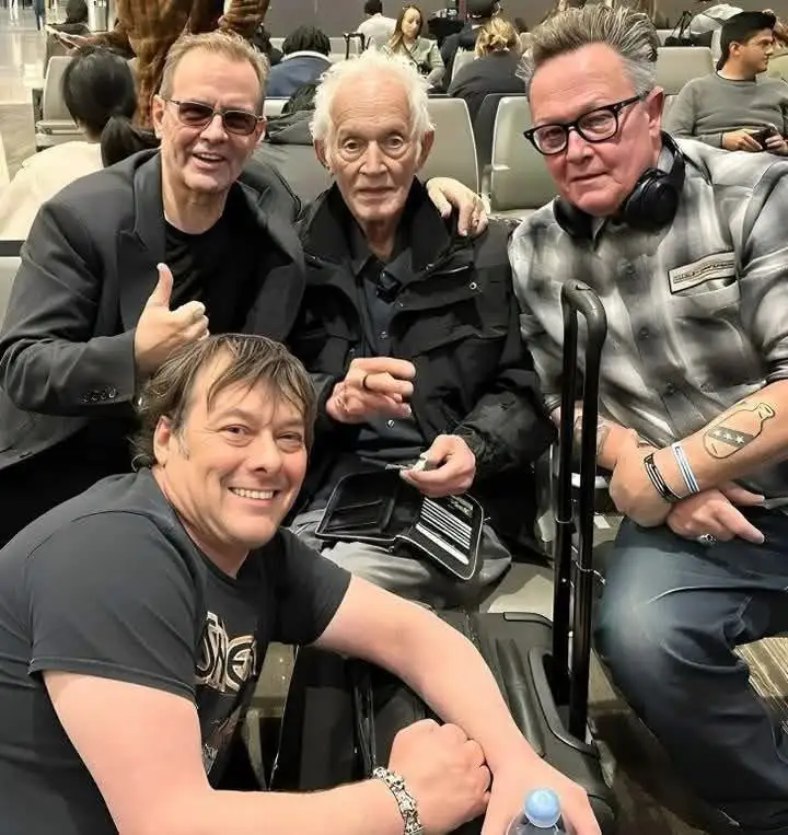 four men, including a smiling man in a black t-shirt in the foreground and three older men behind him in an airport seating…