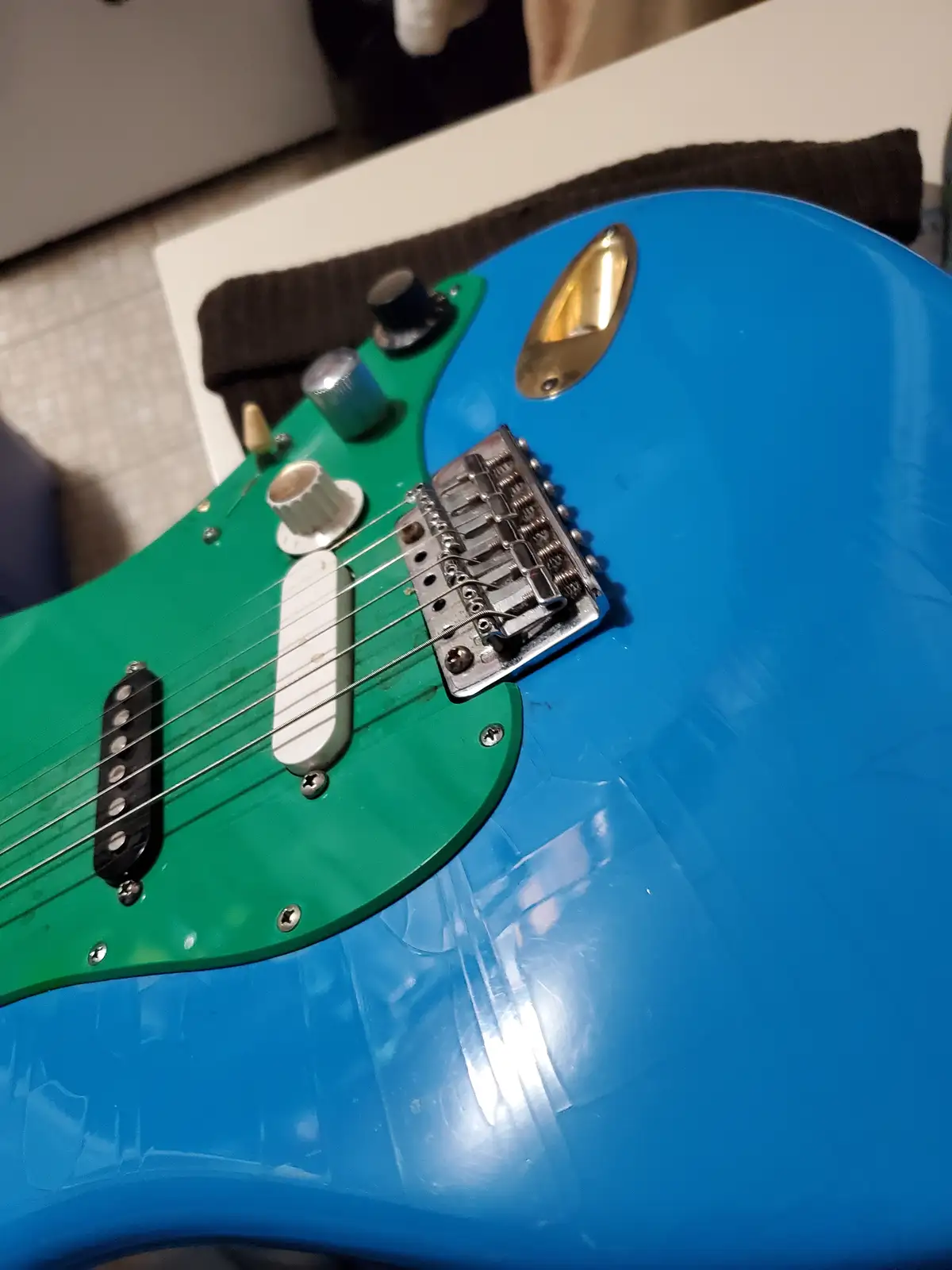 A close-up photo of an electric guitar with a blue body and green pickguard, showing its pickups, knobs, and bridge. The…