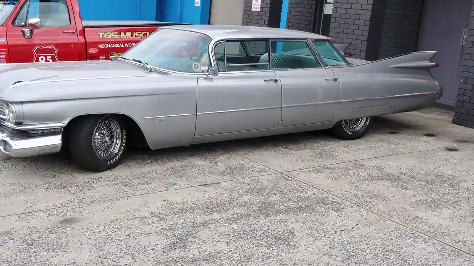 A silver 1960s Cadillac with prominent tail fins and chrome details is parked on a concrete lot, partially obscuring a red…