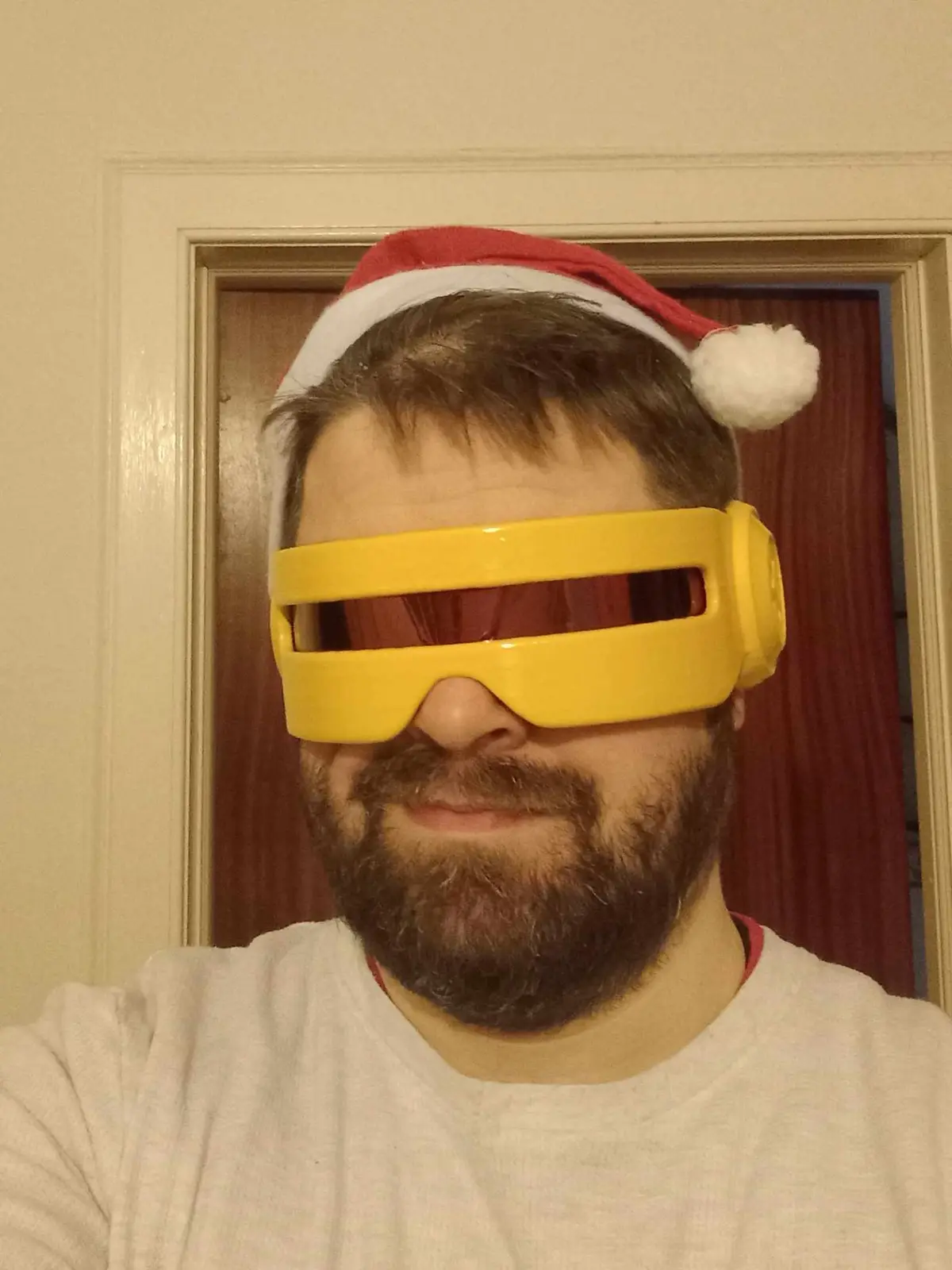 A man with a beard wears a red and white Santa hat and large yellow goggles that cover his eyes, standing in front of a door…