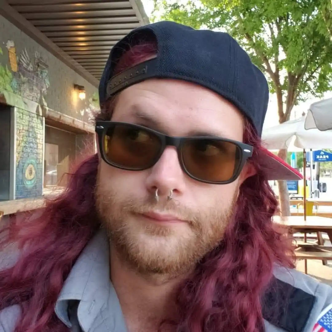 A man with long, wavy reddish-purple hair, a nose ring, and dark sunglasses wears a backward black baseball cap while…
