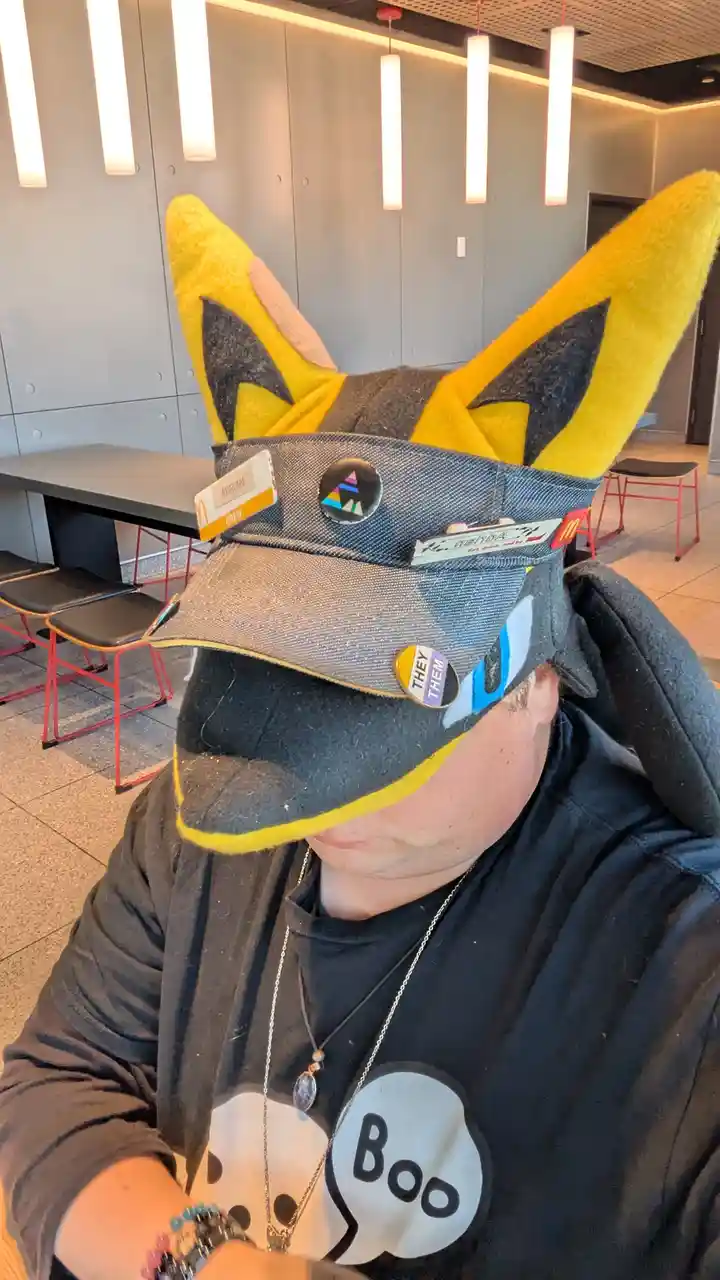 A person wearing a McDonald's crew hat modified with large yellow fox ears and multiple pins, including a 'they/them'…