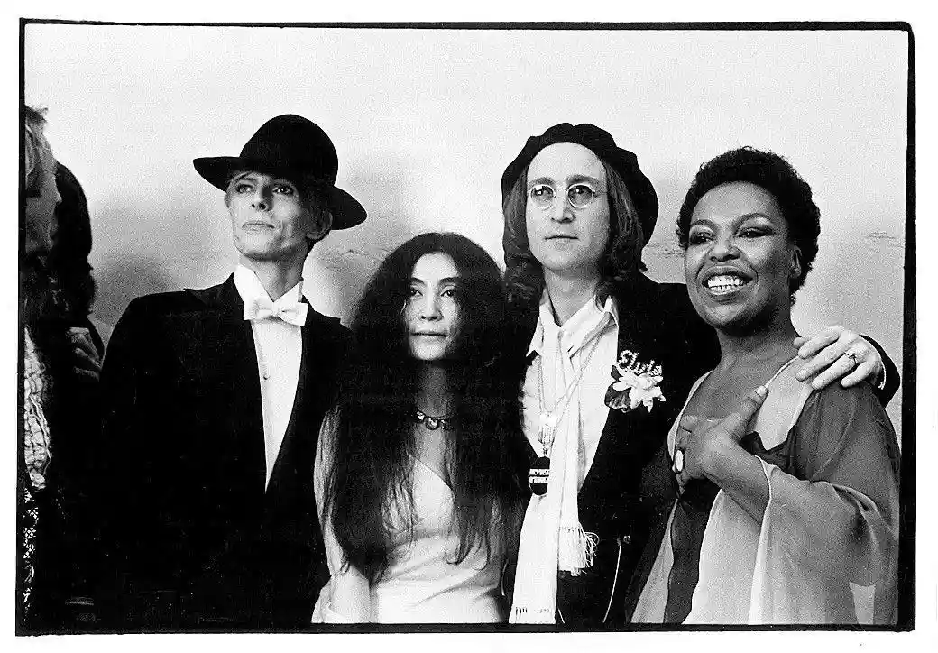 A black-and-white photo of four people posing together; from left to right: a man in a tuxedo and hat, a woman in a white…