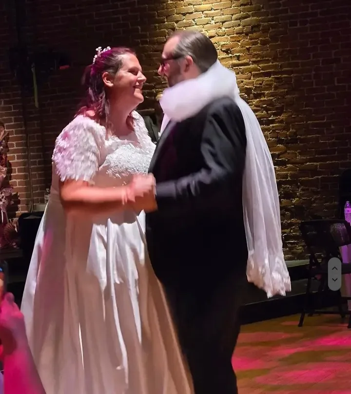 A bride in a white wedding dress with feathered sleeves and a veil dances with a groom wearing a black suit and a large…