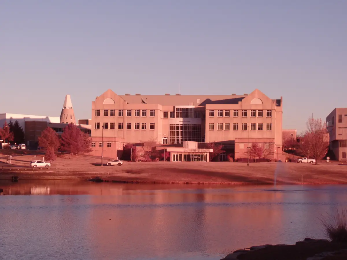 A wide shot of a large, modern institutional building, possibly a university or arts center, viewed across a calm body of…