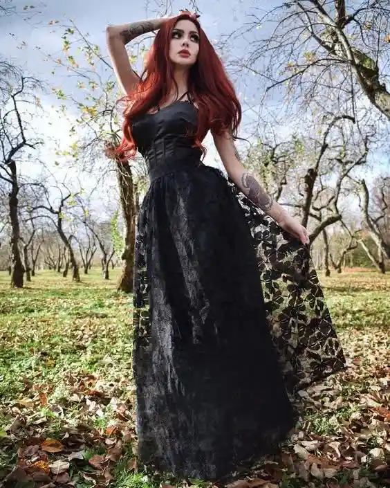 A woman with long red hair and visible tattoos on her arms stands in a leaf-strewn orchard, wearing a black corseted dress…