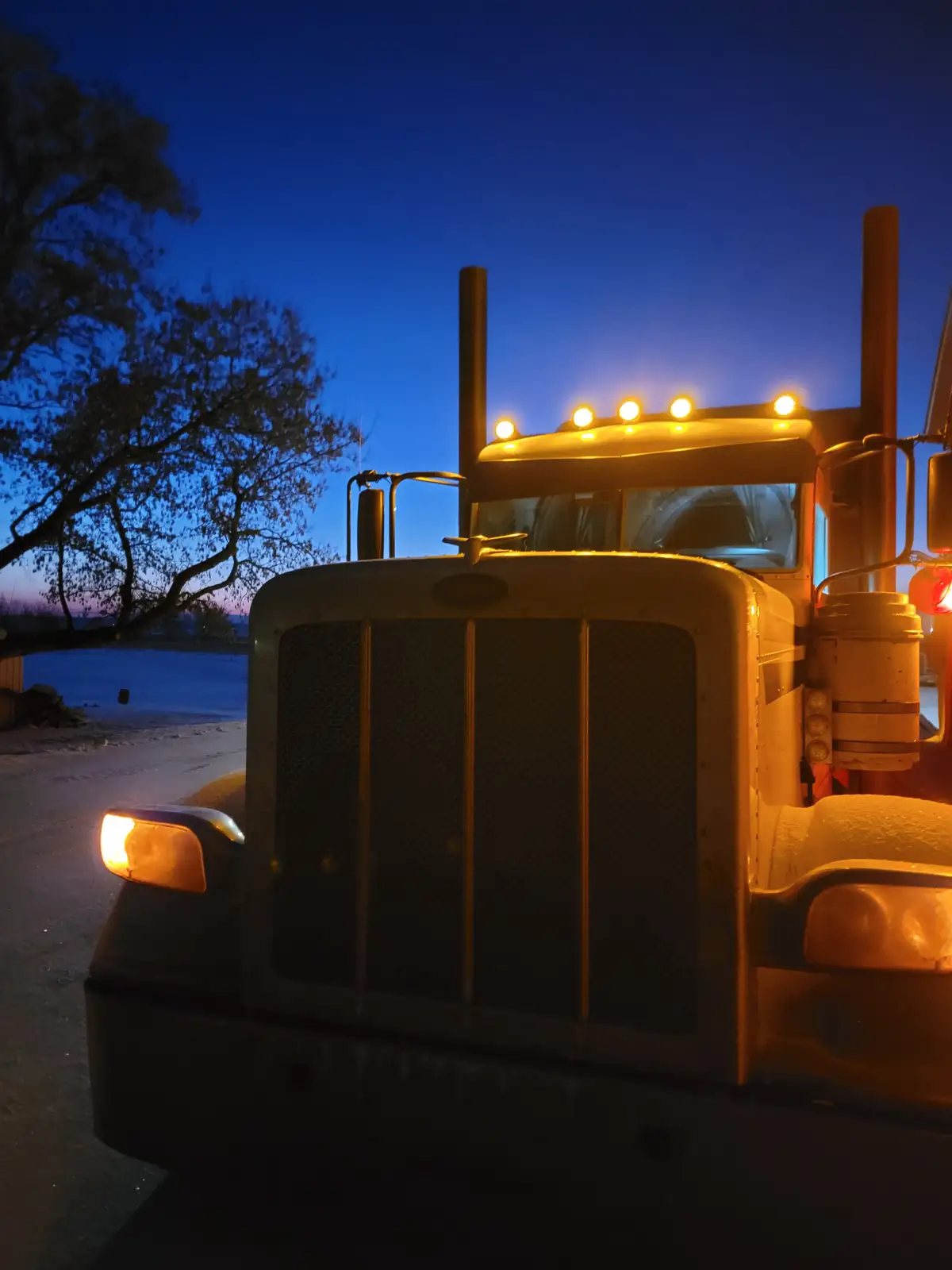 A close-up of a large, illuminated Peterbilt semi-truck at dusk, its headlights and roof lights glowing warmly against a…