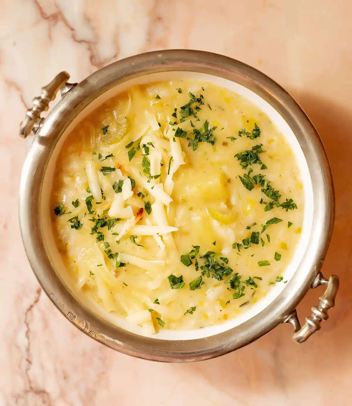 A top-down photo of a creamy, yellowish soup in a silver bowl with ornate handles, garnished with shredded white cheese and…