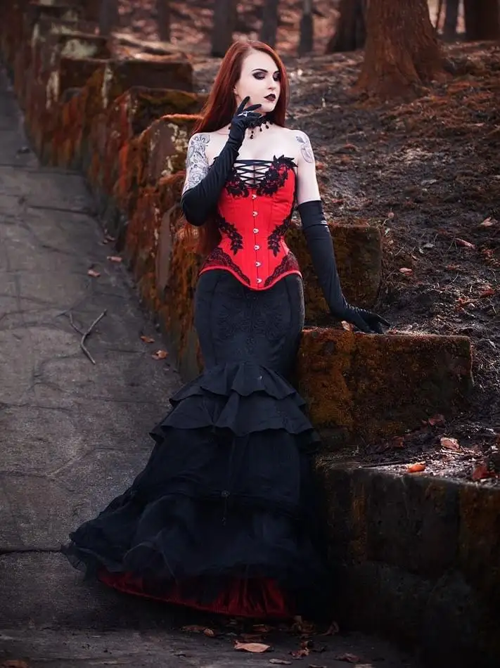 A woman with long red hair, dark makeup, and tattoos poses in a forest path next to mossy stone steps. She wears a red…