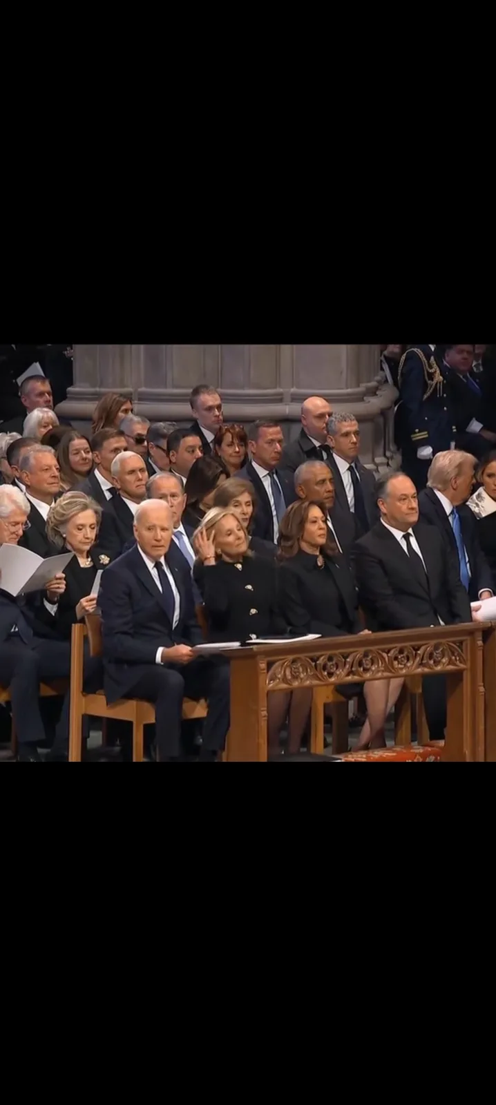 A still image from a formal event shows Joe Biden seated at a wooden table, looking toward Kamala Harris who is seated next…