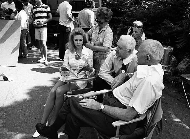 A black-and-white photo shows a woman sitting in a director's chair, looking down at a script with a bored or unimpressed…