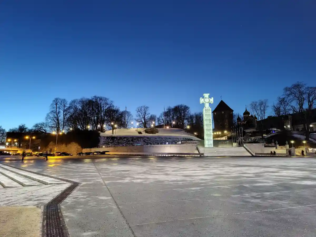 A wide-angle photo of a snow-dusted public square at dusk, featuring a large illuminated cross monument with the text 'EESTI…