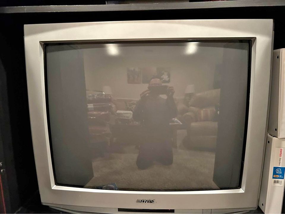 A person is kneeling on a carpeted floor in a living room, taking a photo of themselves with a phone, and their reflection…