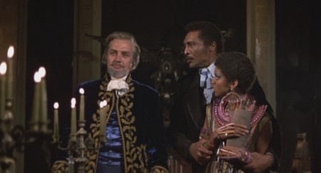 A still from a period drama shows three people in formal 18th-century attire. A white man in a blue velvet coat with gold…