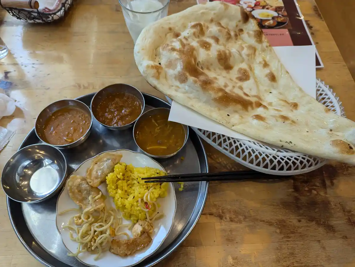 A large, golden-brown naan bread rests on a white basket next to a metal tray holding various Indian dishes including yellow…