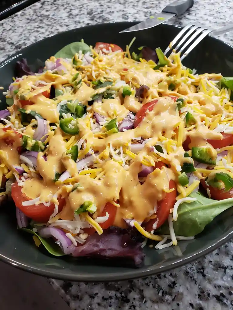A close-up photo of a dark green bowl filled with a salad topped with a thick orange sauce, shredded cheese, chopped…