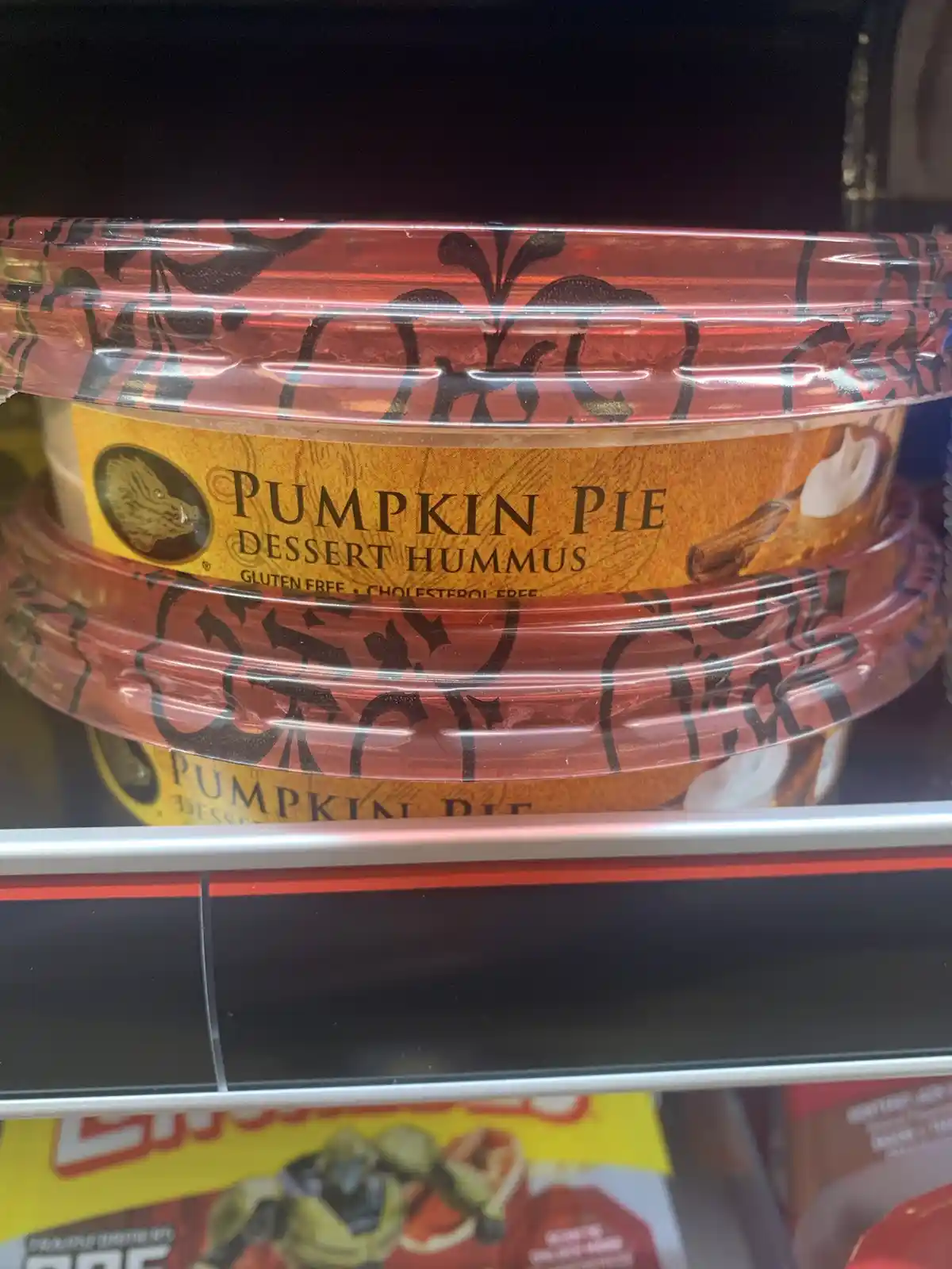 A close-up photo shows stacked containers of 'Pumpkin Pie Dessert Hummus' on a store shelf, with the label clearly visible…