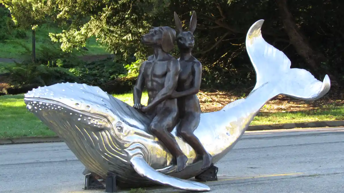 A metal sculpture shows a muscular dog and a kangaroo sitting atop a large, shiny whale. The whale's body is silver and…