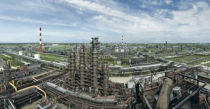 An aerial view of a sprawling, rusted, and abandoned industrial complex with towering pipes, broken structures, and…
