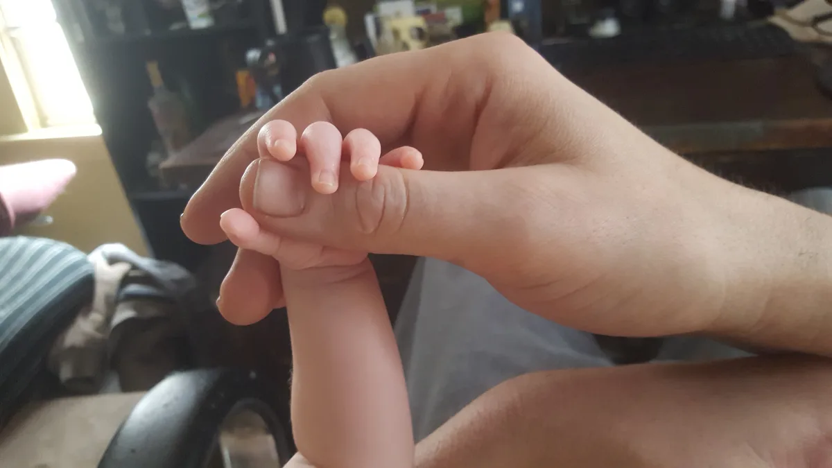 A close-up photo shows an adult's hand gently holding a tiny baby's hand, with the baby's fingers curled around the adult's…