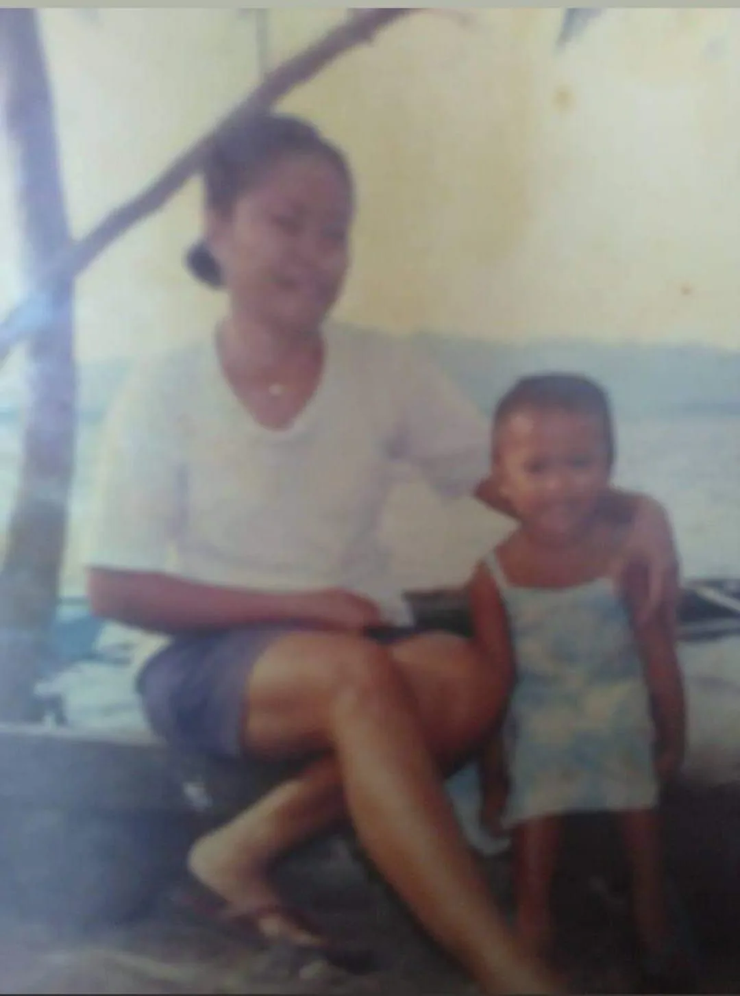 A blurry, low-resolution vintage photograph shows a woman and a small child sitting together outdoors, possibly near water…