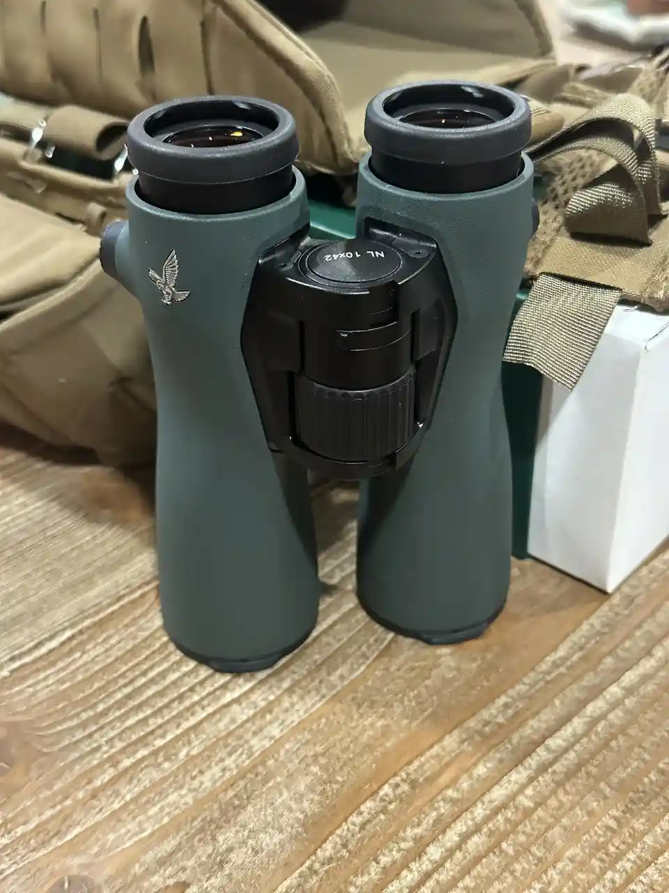 A pair of green binoculars with a silver eagle logo and 'NL 10x42' marking sits on a wooden surface, surrounded by tan…