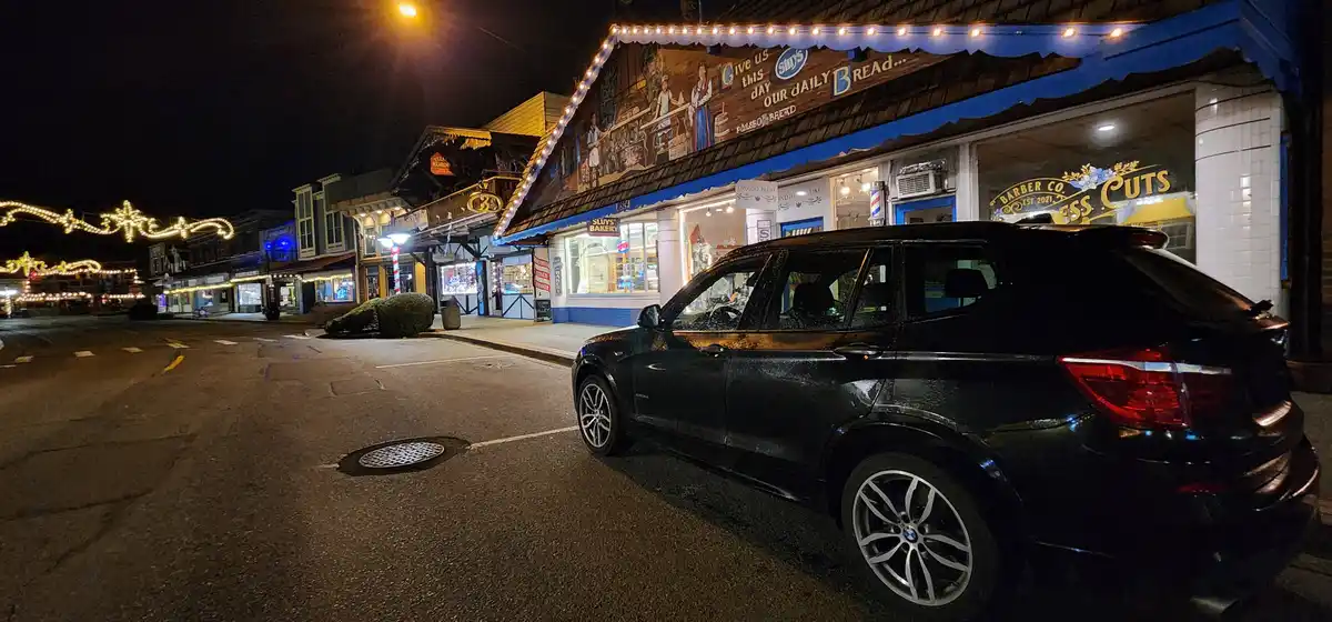 A black BMW SUV is parked on a quiet, illuminated street at night in front of a bakery named 'Sluys Bakery' with a mural…