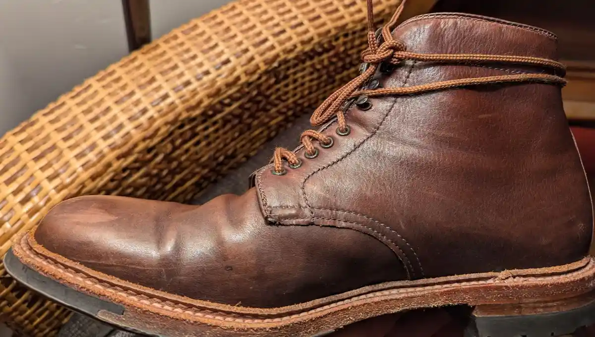 A close-up photo of a well-worn brown leather boot with visible creases and scuffs, resting on a wicker chair. The overlaid…