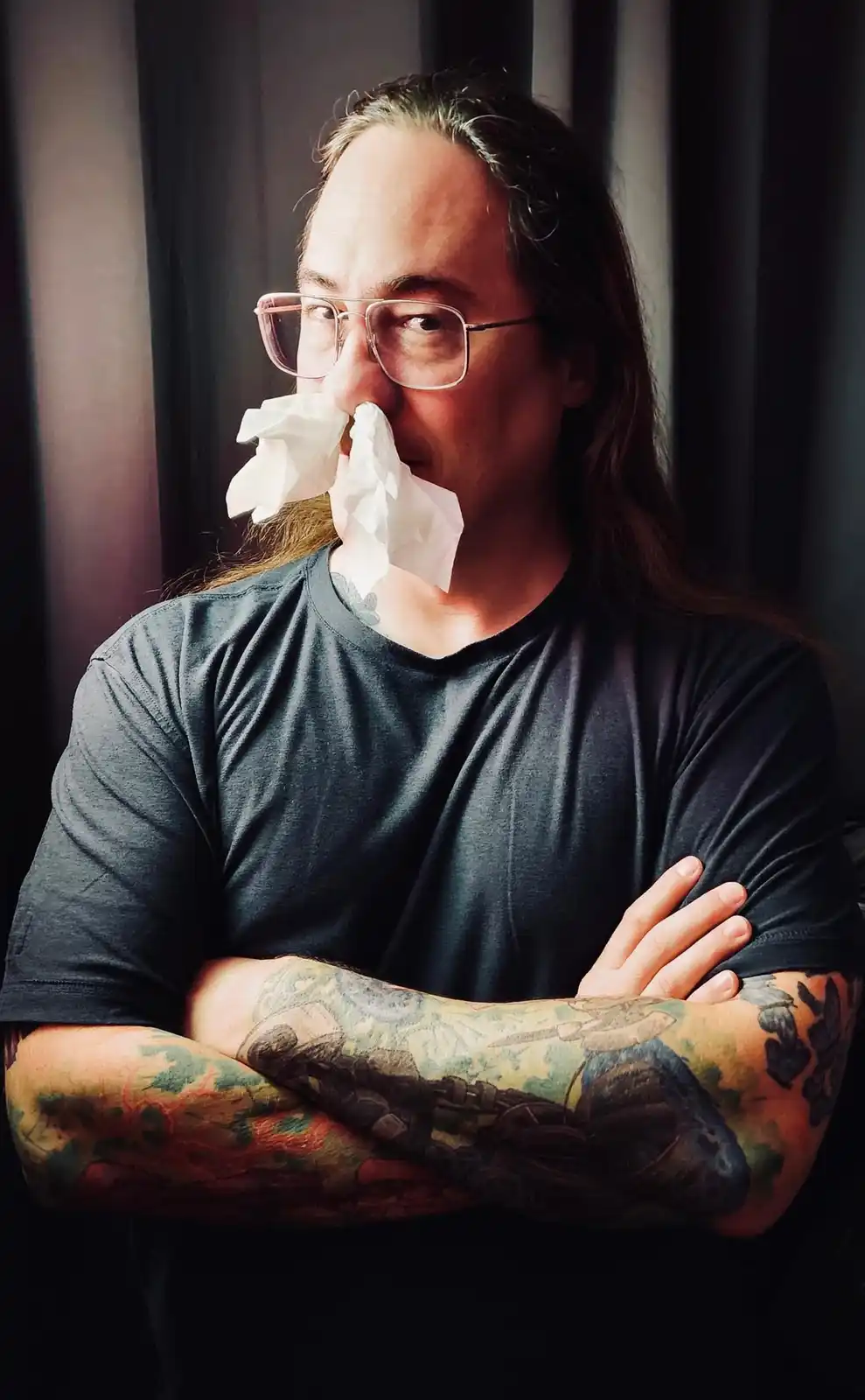A man with long hair, glasses, and heavily tattooed arms stands with his arms crossed, a crumpled white tissue stuffed into…