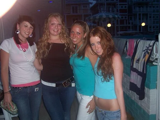 Four young women are posing together on a balcony at night, smiling for the camera. Behind them, towels hang on a railing…