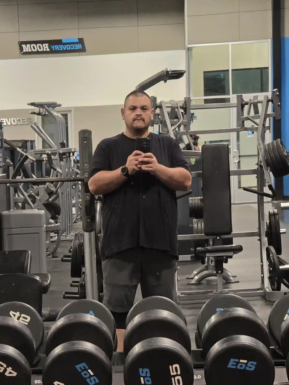 A man with a mustache and earbuds stands in a gym mirror selfie, holding a phone, surrounded by weight machines and…