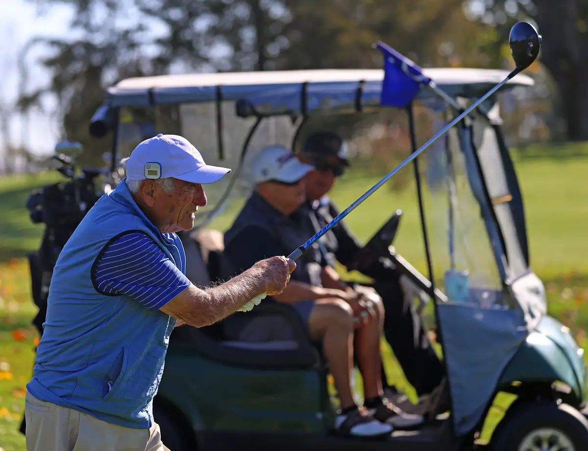 An older man in a blue vest and white cap is mid-swing with a golf club on a sunny course, with two other men in a golf cart…