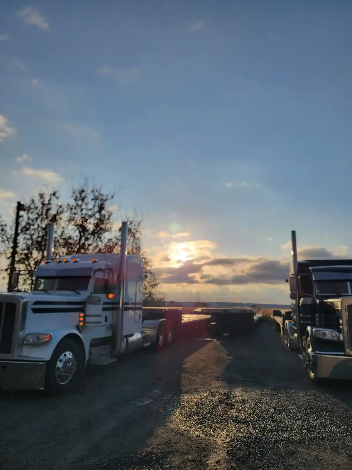 Two large semi-trucks are parked on a gravel lot facing each other with the sun setting behind them, casting long shadows…