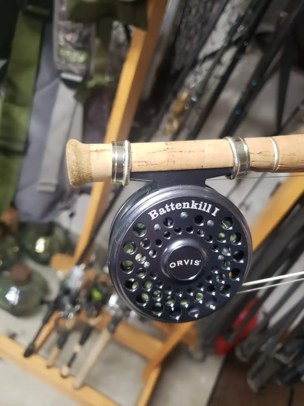 A close-up photo of an Orvis Battenkill I fly fishing reel attached to a cork-handled rod, with other fishing gear blurred…