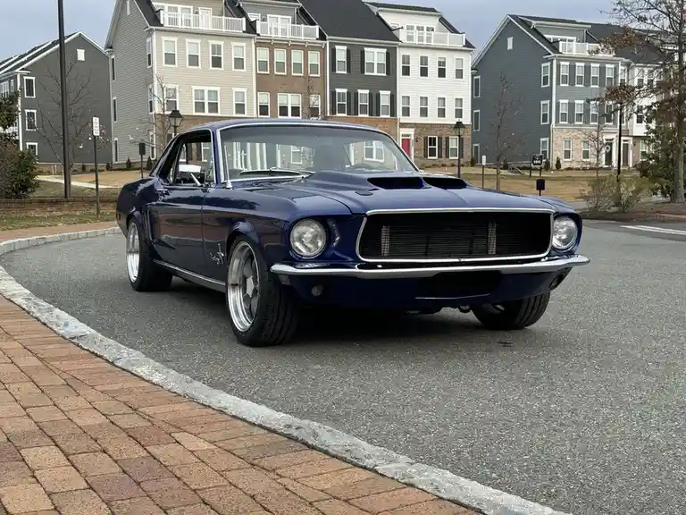 A glossy blue 1960s Ford Mustang with chrome trim and modern silver wheels is parked on a residential street, facing the…
