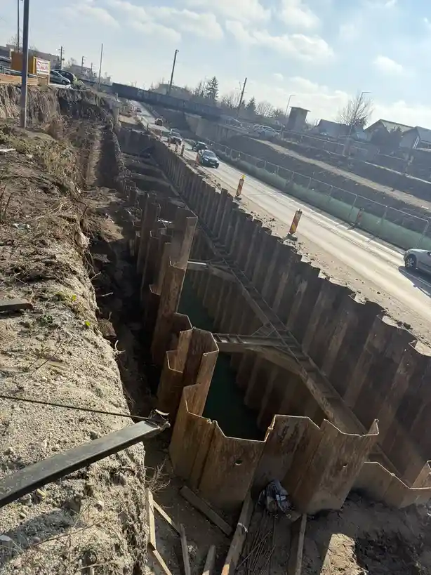 A photo of a deep construction trench with wooden sheeting and water inside, running parallel to a road with cars passing by…