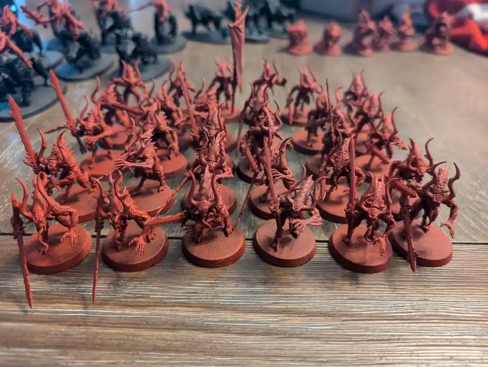 A collection of miniature fantasy figurines, mostly painted a uniform red, are arranged on a wooden surface. Some are in the…