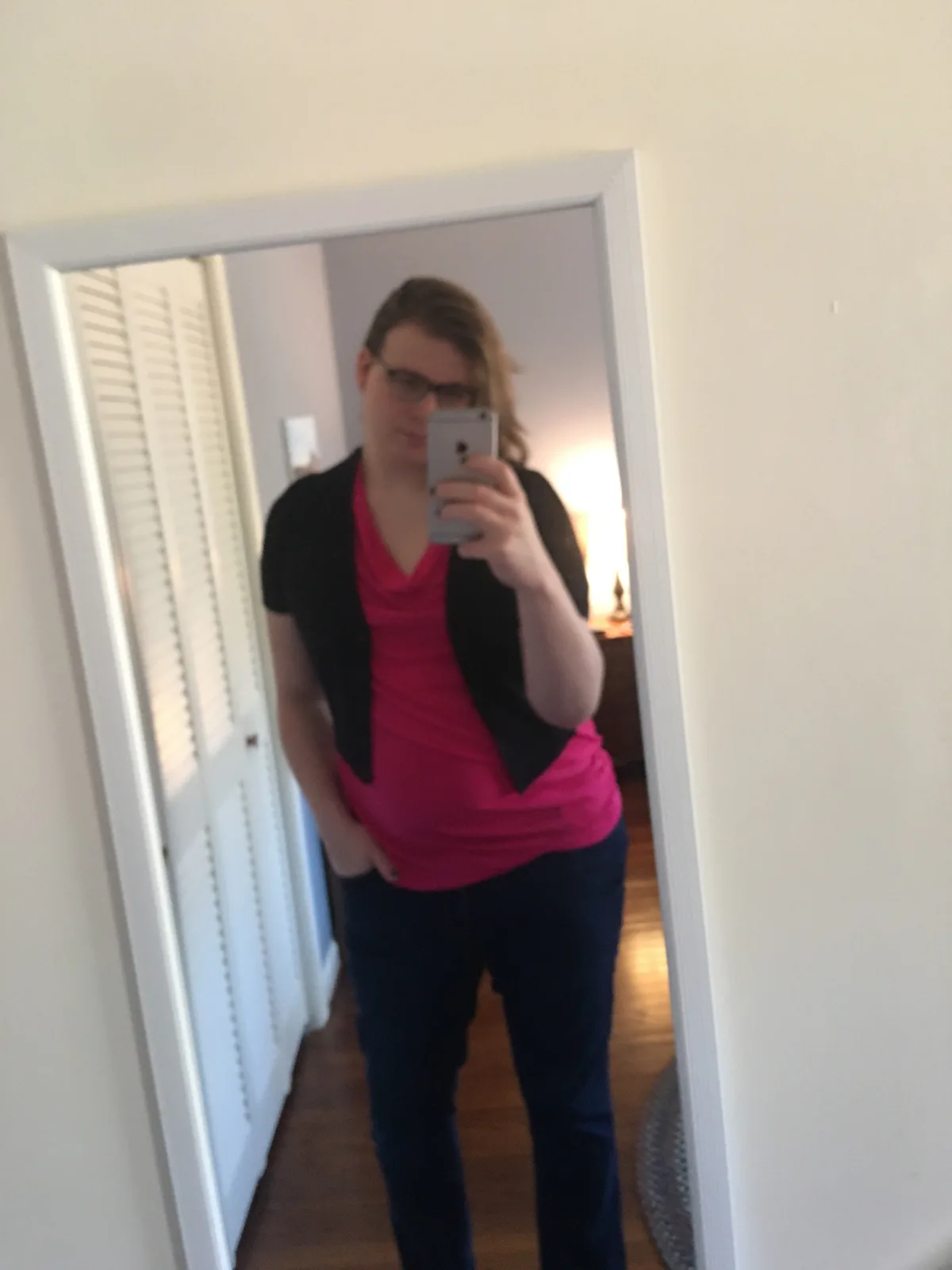 A person with glasses and long hair takes a mirror selfie in a room with white walls and wooden floors, wearing a pink shirt…