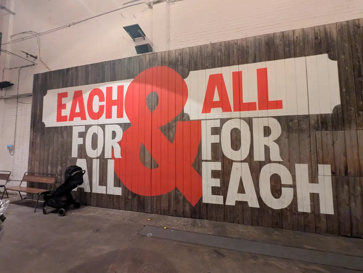 A large wooden wall mural displays the phrase 'EACH & ALL FOR ALL FOR EACH' in bold red and white letters, with a large red…