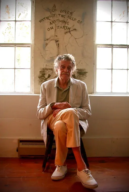 An elderly man with gray hair and a mustache sits calmly on a wooden stool between two large windows in a sunlit room. He…