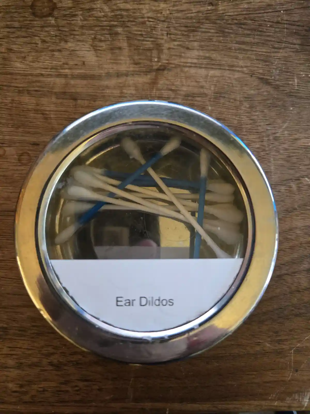 A top-down view of a metal jar with a white label reading 'Ear Dildos' sitting on a wooden surface. Inside the jar are…