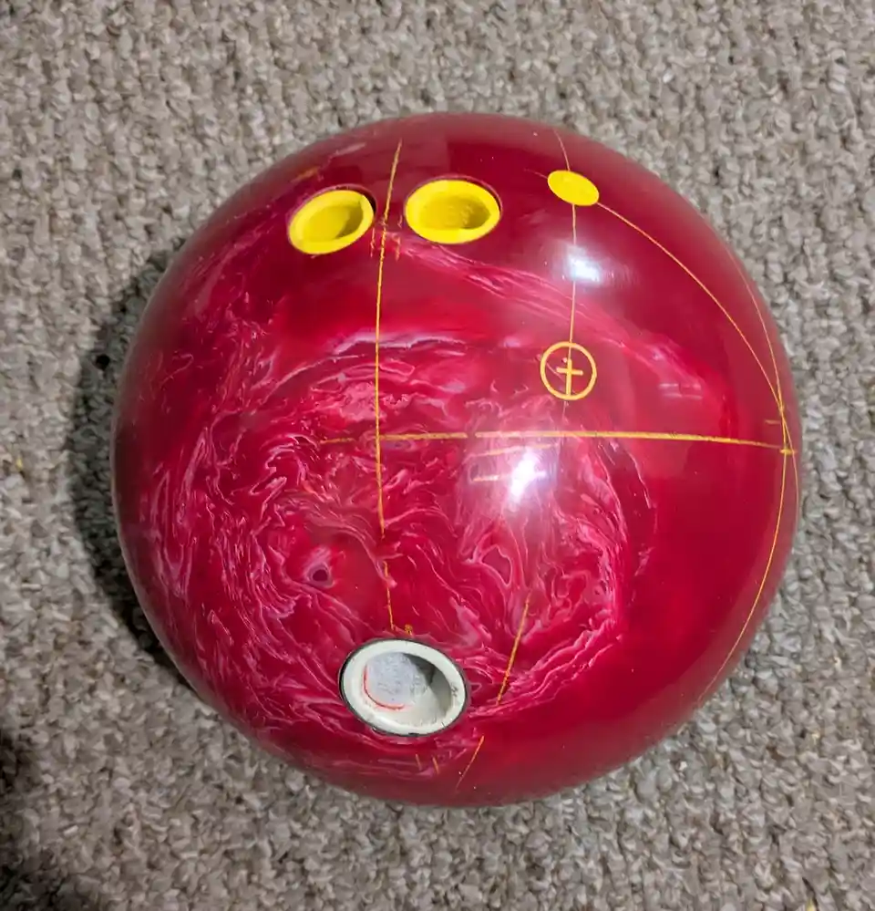 A red marbled bowling ball with yellow finger holes and a white thumb hole rests on a beige carpet. Yellow lines and a…