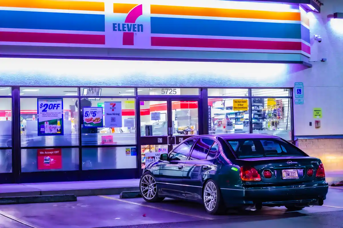 A dark green Lexus sedan with aftermarket wheels is parked in front of a brightly lit 7-Eleven store at night. The store's…