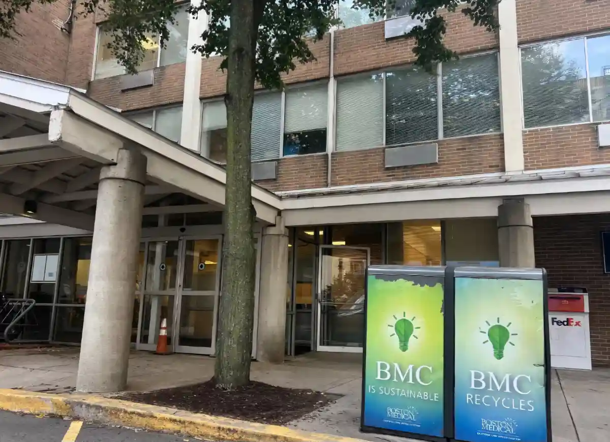 An exterior shot of a brick building with glass doors and windows, featuring two adjacent green and blue recycling bins in…