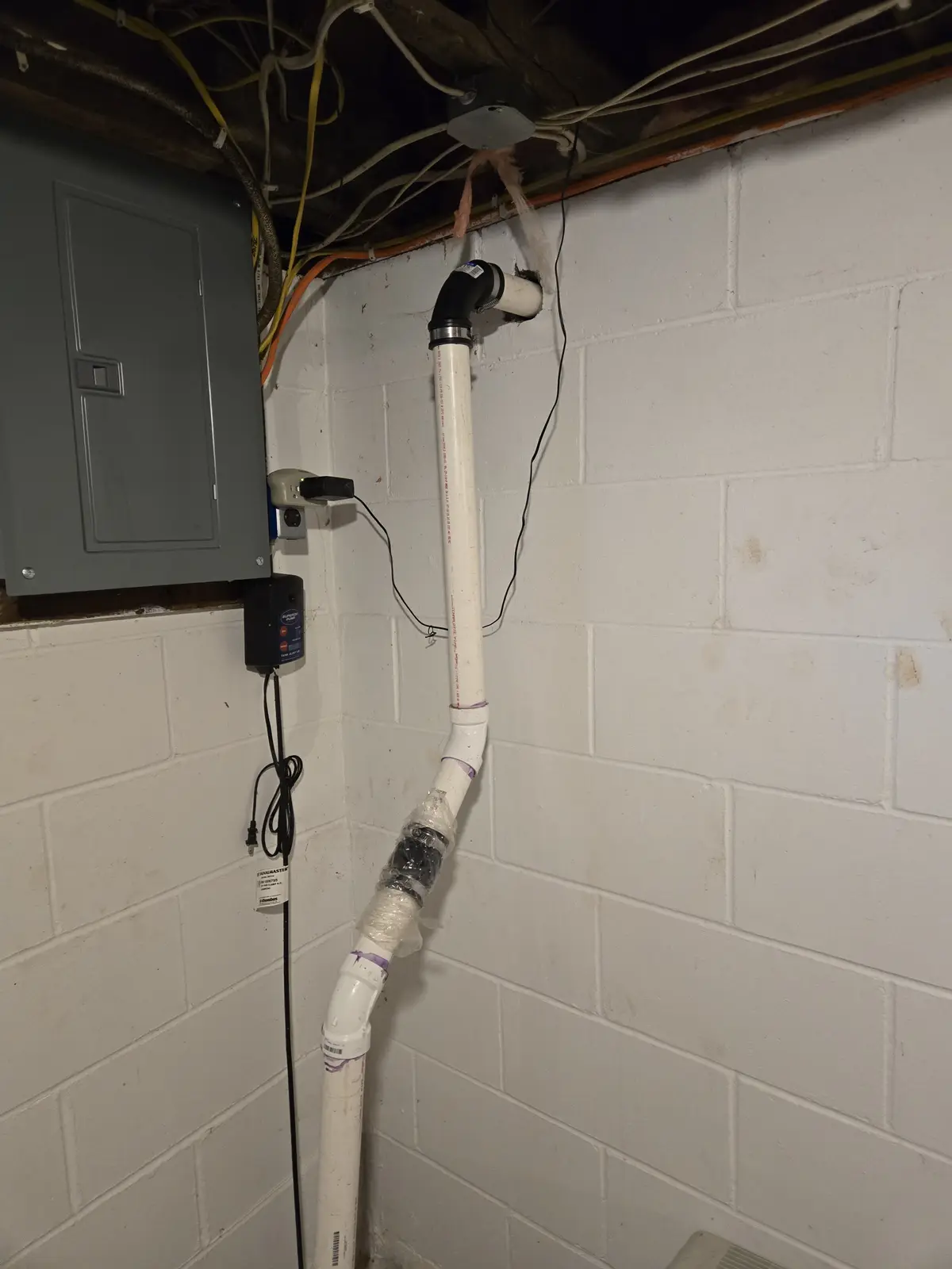 A basement corner with white cinder block walls, a grey electrical panel, and a white PVC pipe running vertically. The pipe…