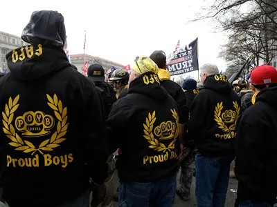 A group of men viewed from behind, wearing black hoodies with gold 'Proud Boys' logos and laurel wreaths. Some have 'Proud…