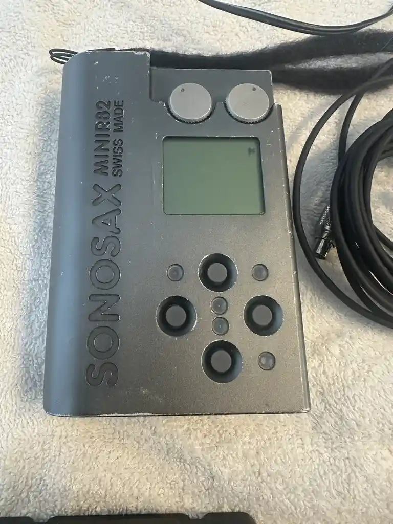 A gray Sonosax Minir32 Swiss Made device with a small screen, two dials, and multiple buttons sits on a white towel next to…