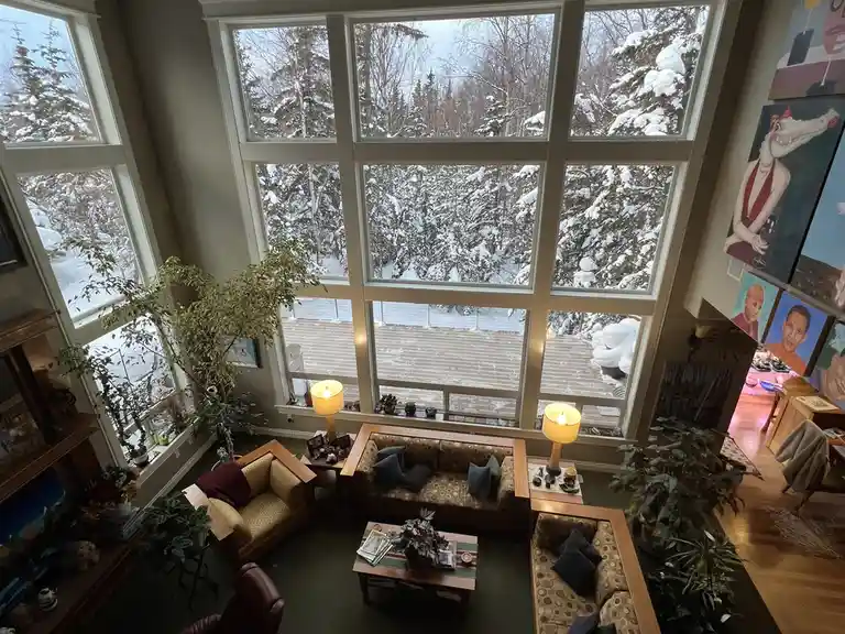An overhead photo of a cozy, well-furnished living room with large windows overlooking a snowy forest. The room contains…
