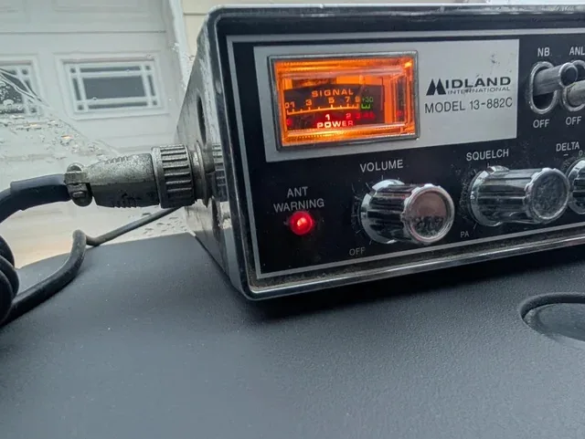 A close-up of a Midland International Model 13-882C handheld radio with a glowing orange display showing 'SIGNAL 1 2 7 8 9…