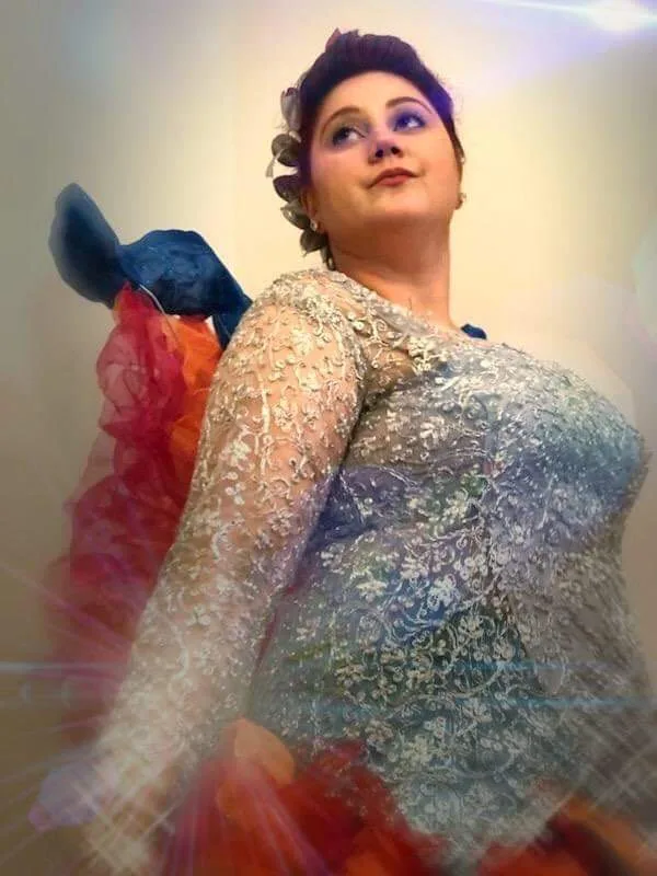A woman in an ornate, sparkly blue and silver dress with a colorful, layered skirt looks upward with a confident, slightly…
